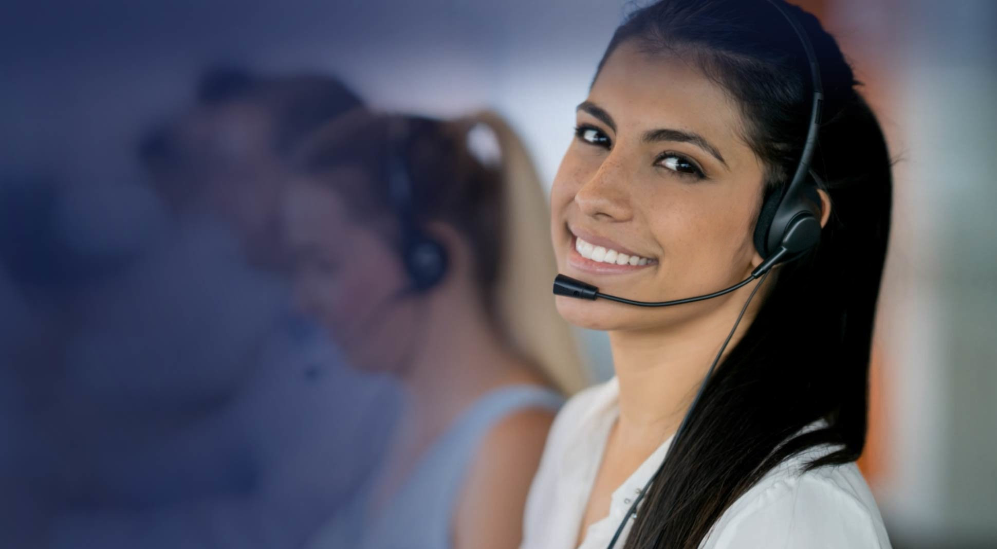 Mujer joven con auriculares y micrófono de atención al cliente, trabajando en un centro de llamadas, con fondo desenfocado de otros operadores.